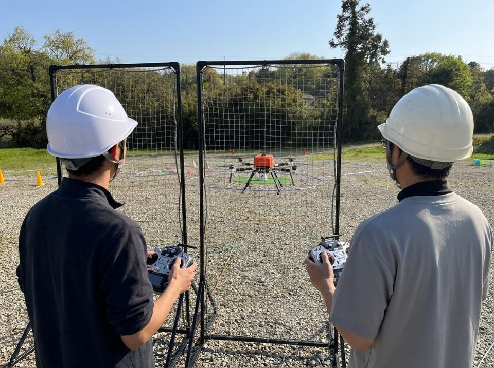 防護ネットの後ろでヘルメットを着用した受講生と講師が送信機を持ち、自社製作機を操縦する実地試験対策のイメージ