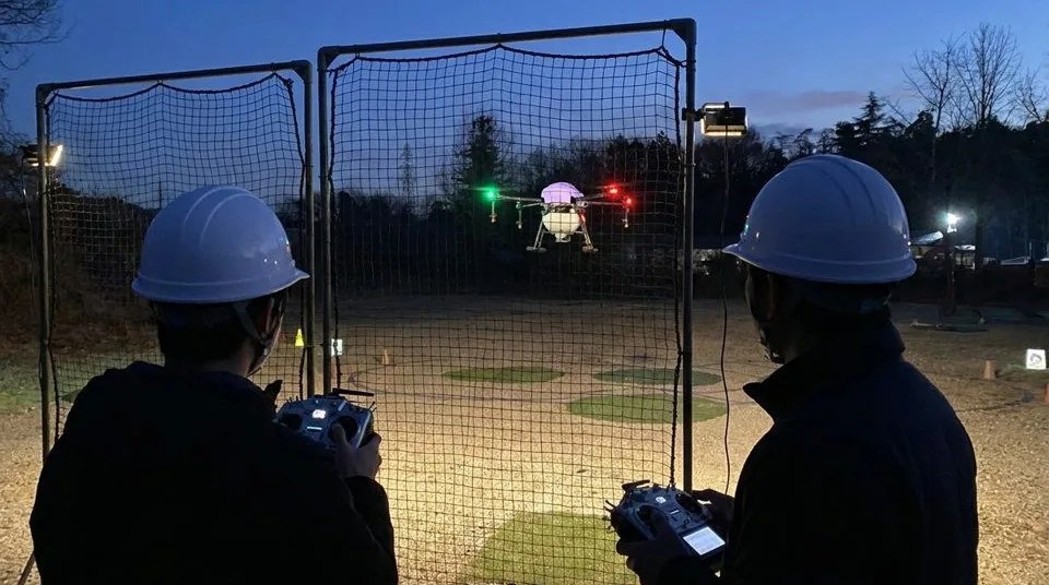 夕方の飛行場で、防護ネットの後ろから受講生がドローンを操縦する実地講習の様子